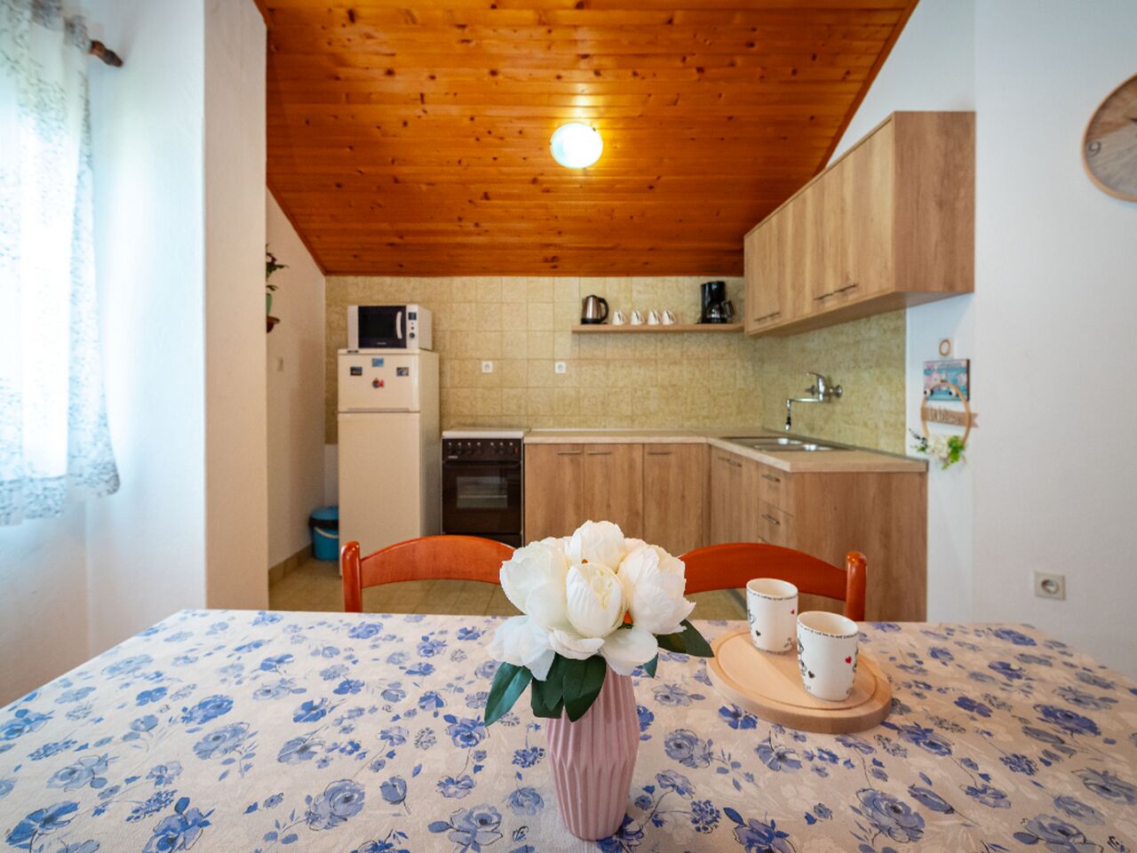A dining table with a floral tablecloth, a vase with white flowers, and two mugs on a tray.