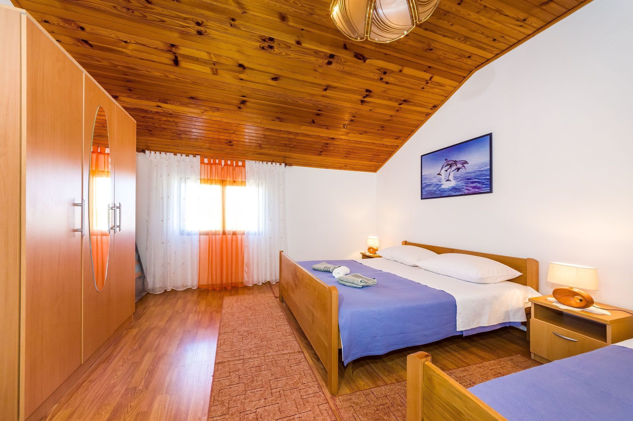 Una camera da letto con soffitto in legno, un letto con copriletto blu, un comodino con una lampada e una finestra con tende.