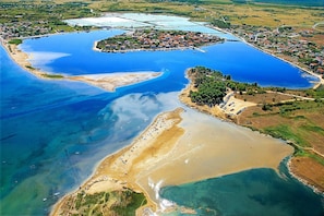 An aerial view of a coastal area with a large body of water, a small island with buildings, and a beach with people and boats.