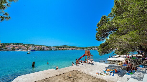 Um parquinho à beira-mar com escorrega e área de areia, com vista para um mar azul e límpido, com barcos e edifícios ao fundo.