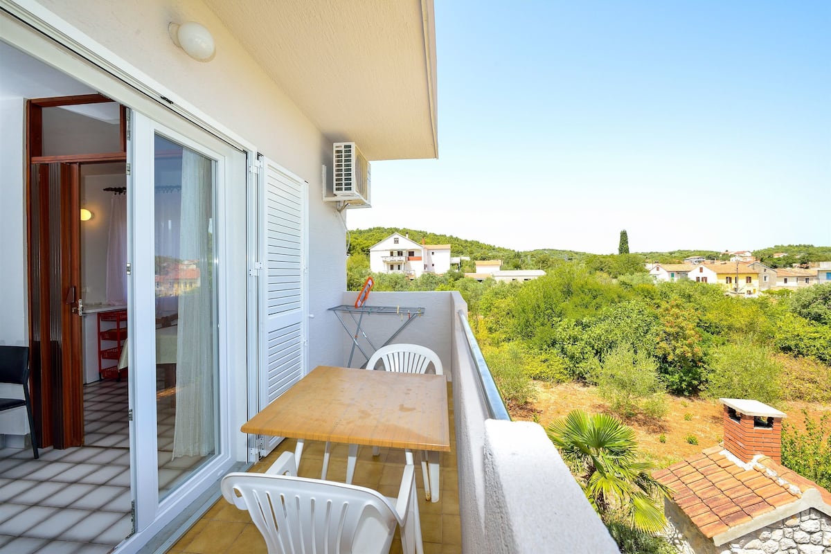 A balcony with a table and chairs, a sliding glass door, and a view of a hillside with buildings.
