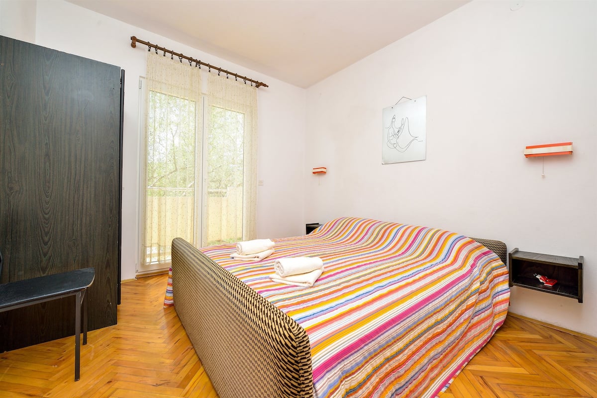 A bedroom with a striped bedspread, a bench, a wardrobe, and a window with curtains.
