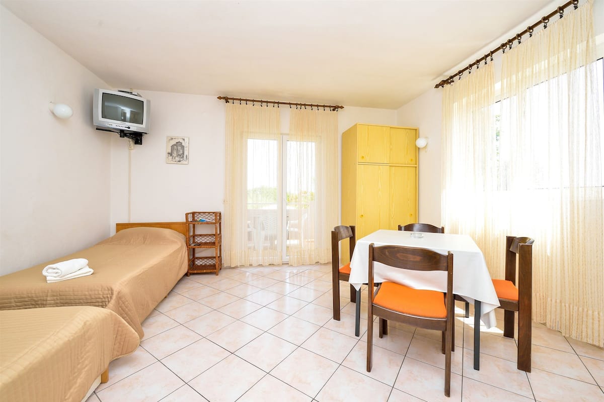 A room with two single beds, a dining table with four chairs, a television mounted on the wall, and a window with curtains.