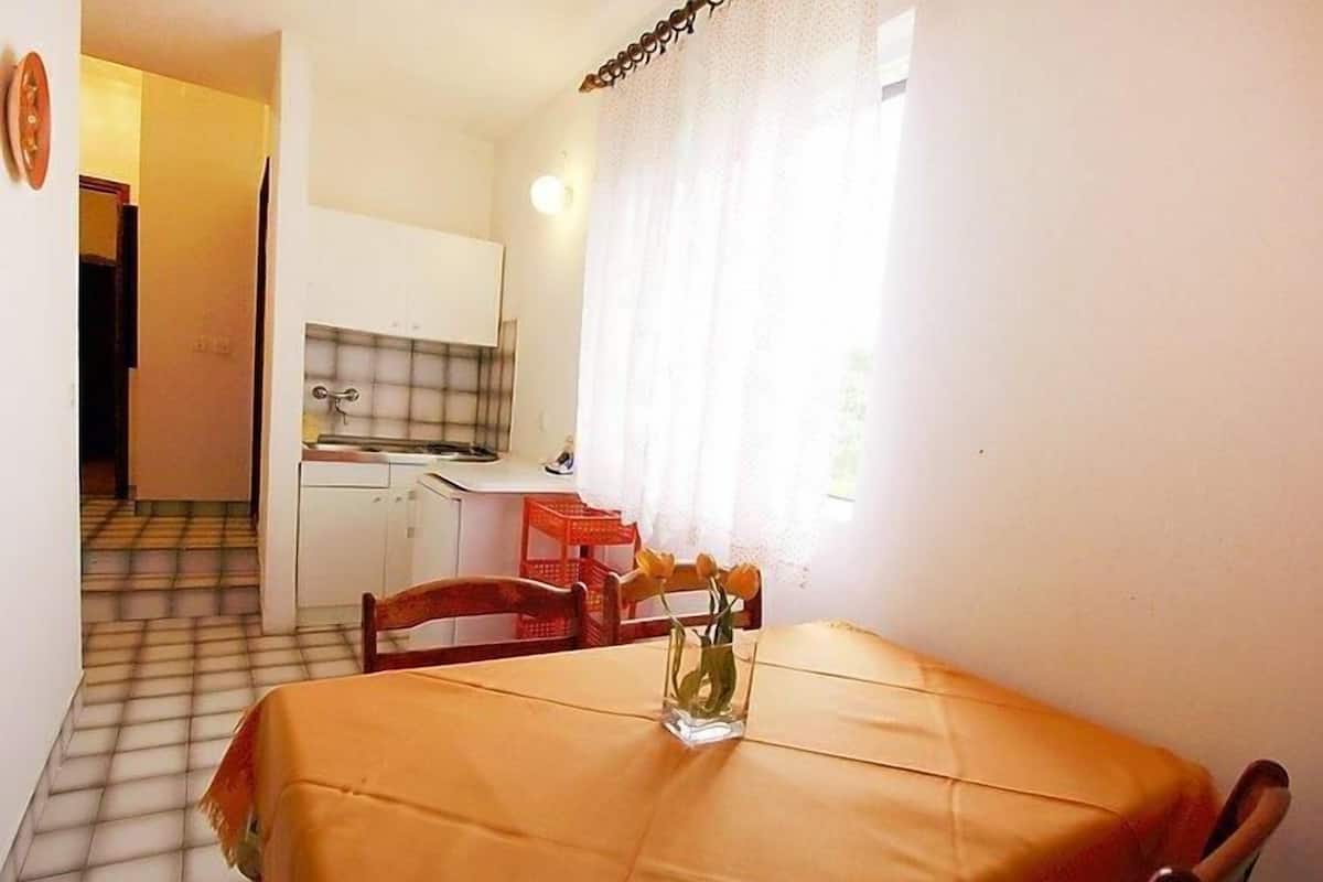A small kitchen with a tiled backsplash, a sink, and a window with curtains. There is a dining table with a tablecloth and a vase with flowers.