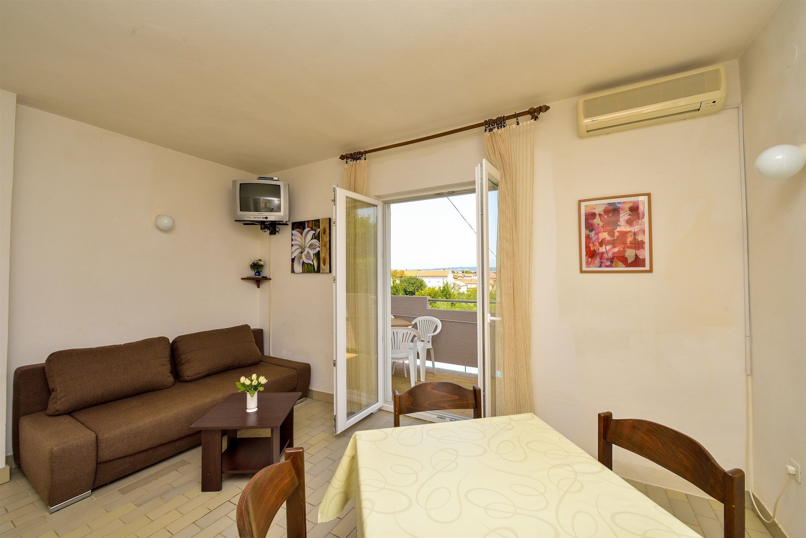 A living room with a sofa, a table, and a TV. There is a balcony with chairs and a table.