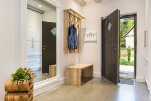 A hallway with a wooden bench, a coat rack with a blue jacket, a wooden planter with flowers, and a door leading outside. - AS1A - Old barn (AS) at c a l l s e n - appartements (Ummanz)