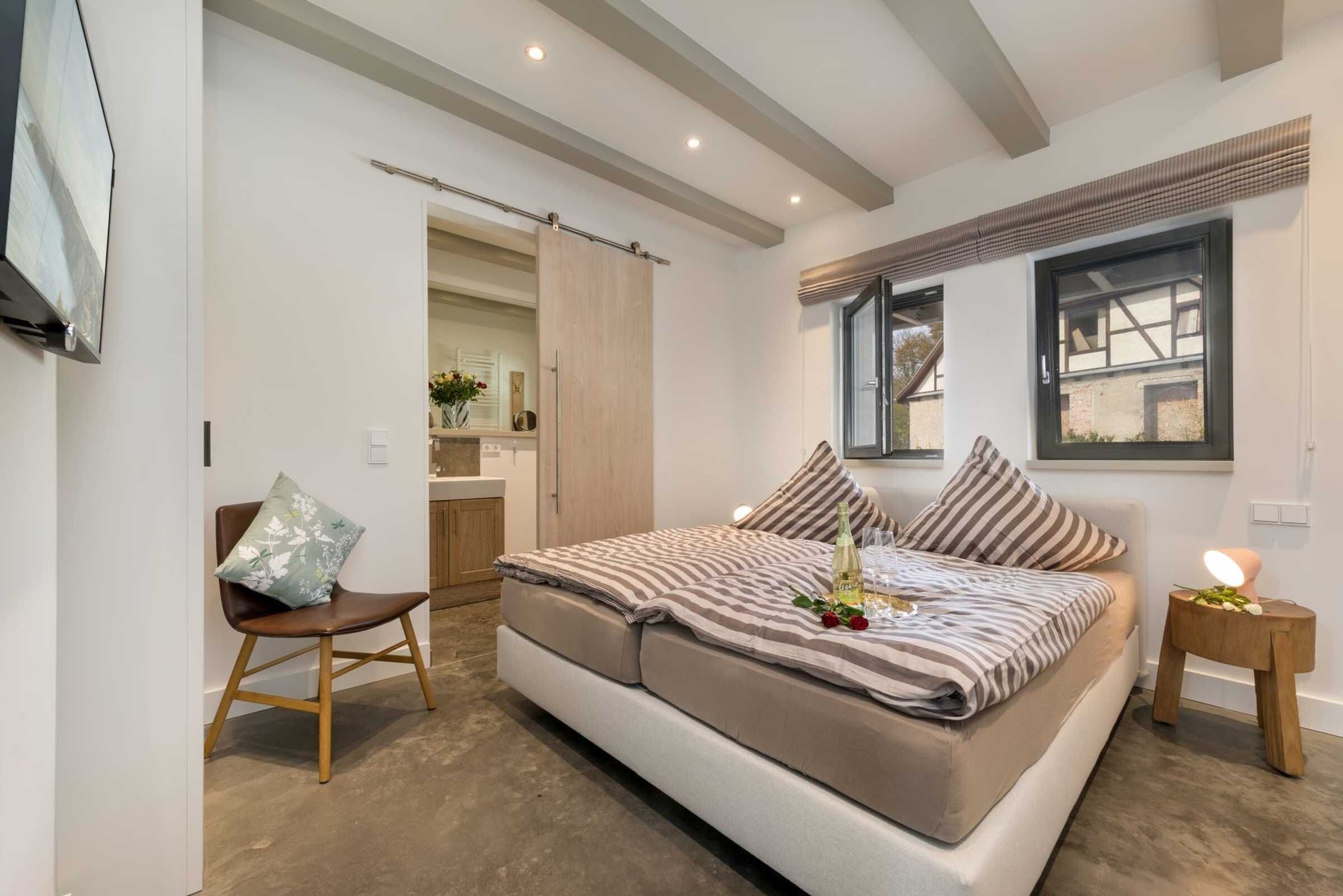 Un dormitorio con cama, una silla, una mesita de noche, una ventana y una puerta.
