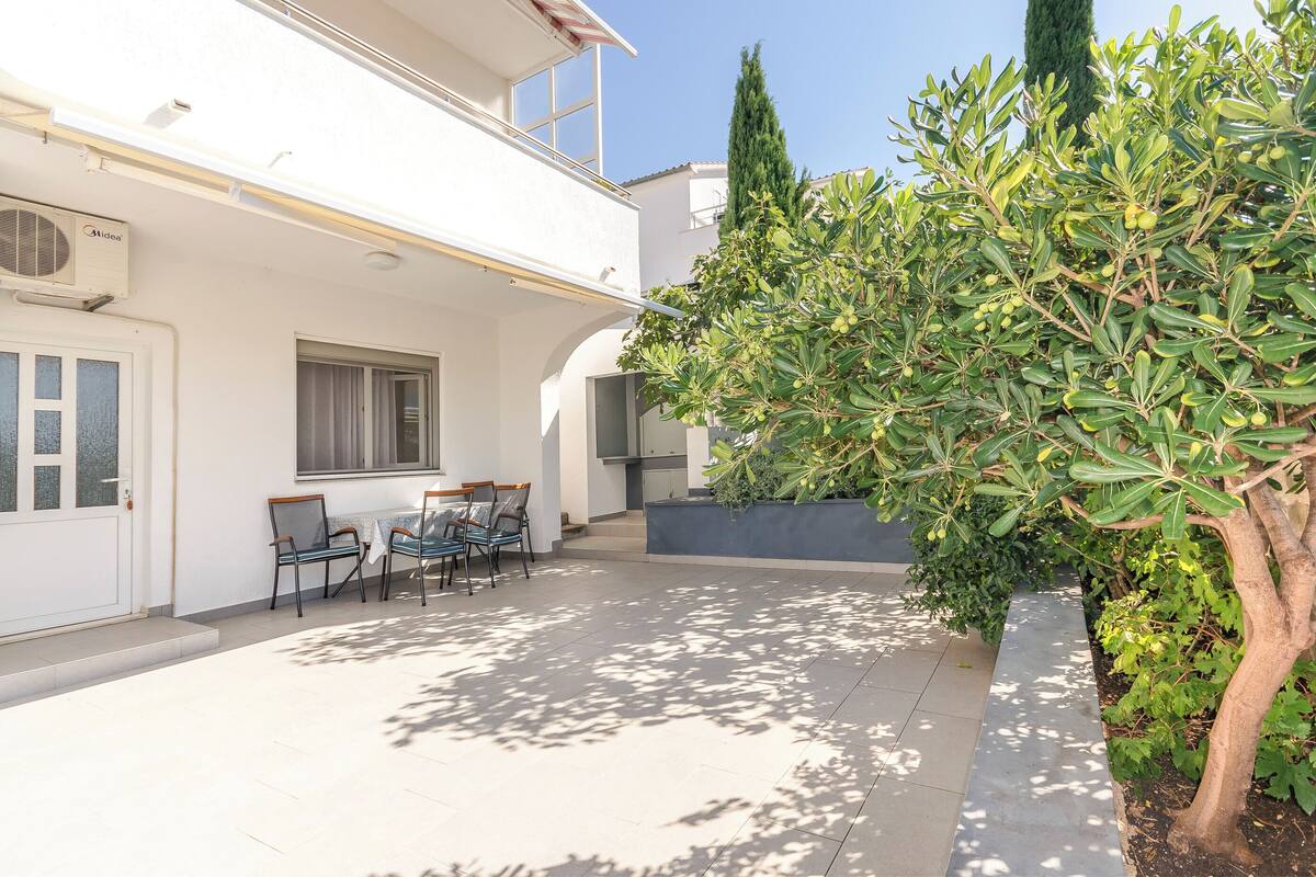 A white building with a patio area featuring a table and chairs, a small garden with a tree, and a clear sky.