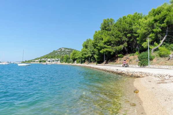 A coastal scene with clear water, boats, and a road with trees and a hill in the background.