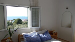 Ένα υπνοδωμάτιο με ένα κρεβάτι, ένα μικρό κομοδίνο, ένα φωτιστικό, ένα παράθυρο με θέα στη θάλασσα και τα σπίτια, και έναν καθρέφτη στον τοίχο.