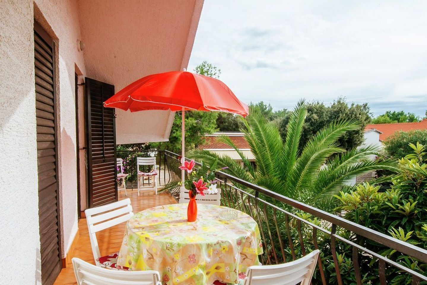 A balcony with a table and chairs, a red umbrella, and lush greenery in the background.