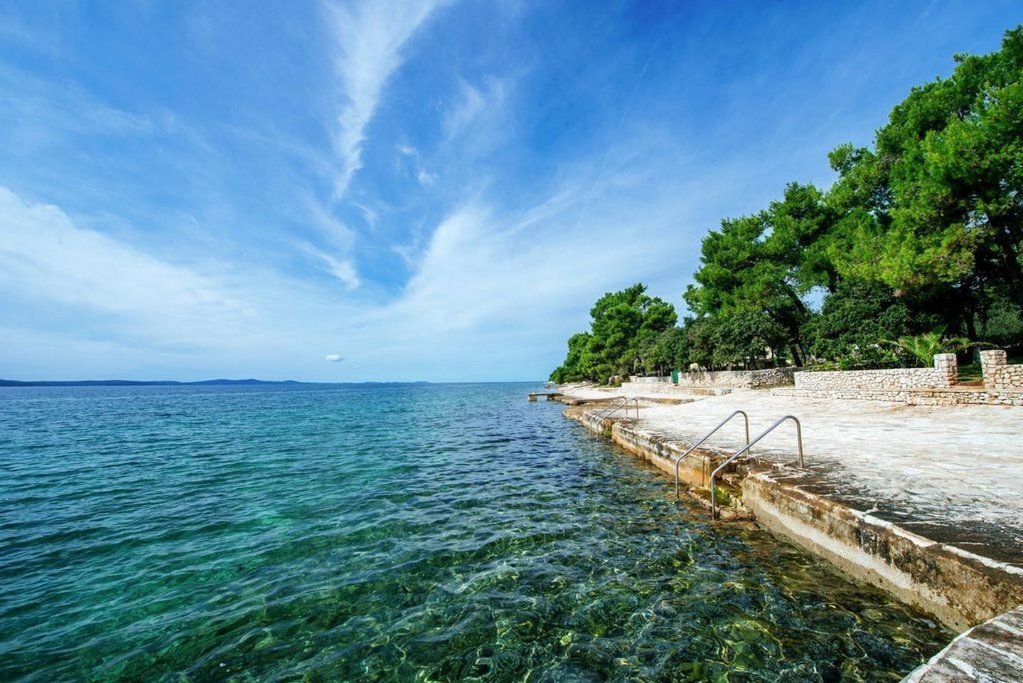 A clear blue sea with a rocky pier and trees.