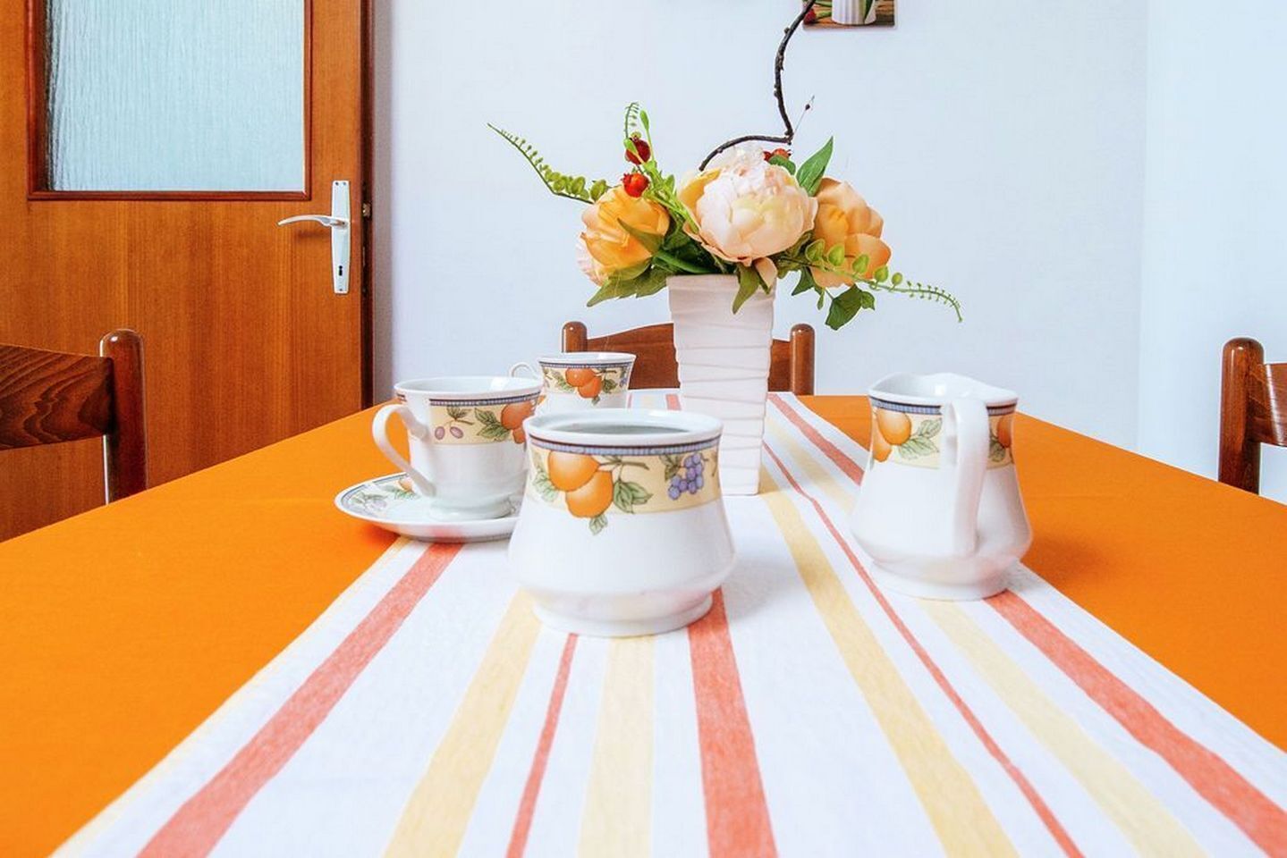 A dining table with striped tablecloth, white ceramic tea set, and a vase with flowers.