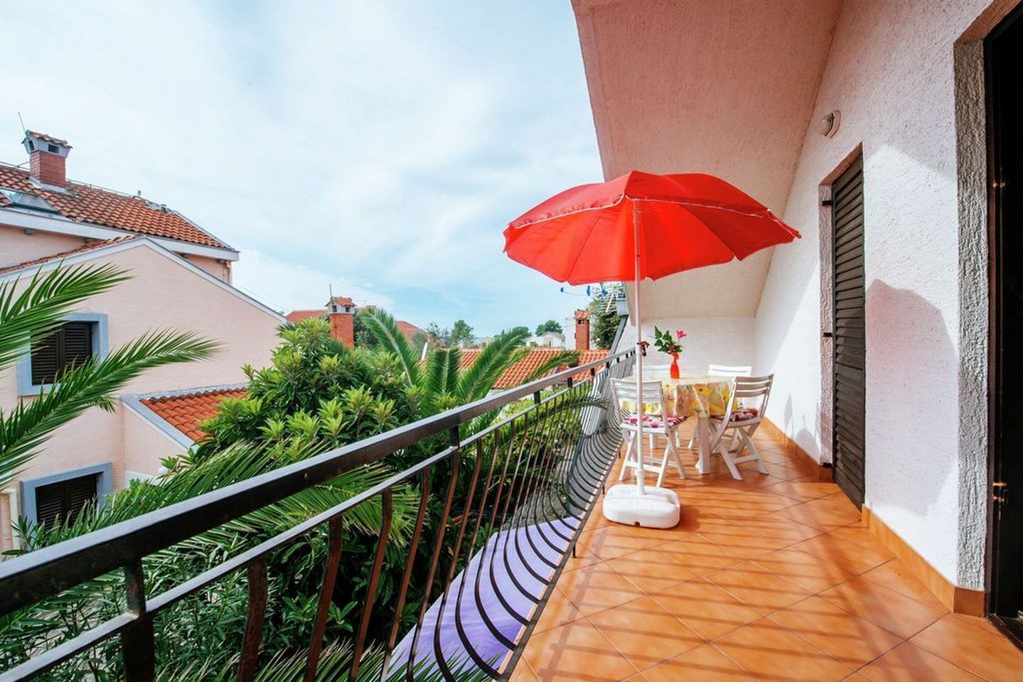 Ein Balkon mit einem roten Sonnenschirm, weißen Stühlen und einem Tisch mit einer Vase.