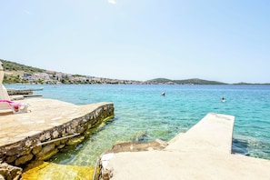A stone pier extending into clear blue water with a view of a hillside village in the background.