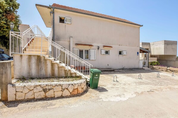 A two-story building with a stone staircase leading to the entrance, a green trash bin, and a concrete area with three metal faucets. - Apartments Apartmani Bebe (Razanj)