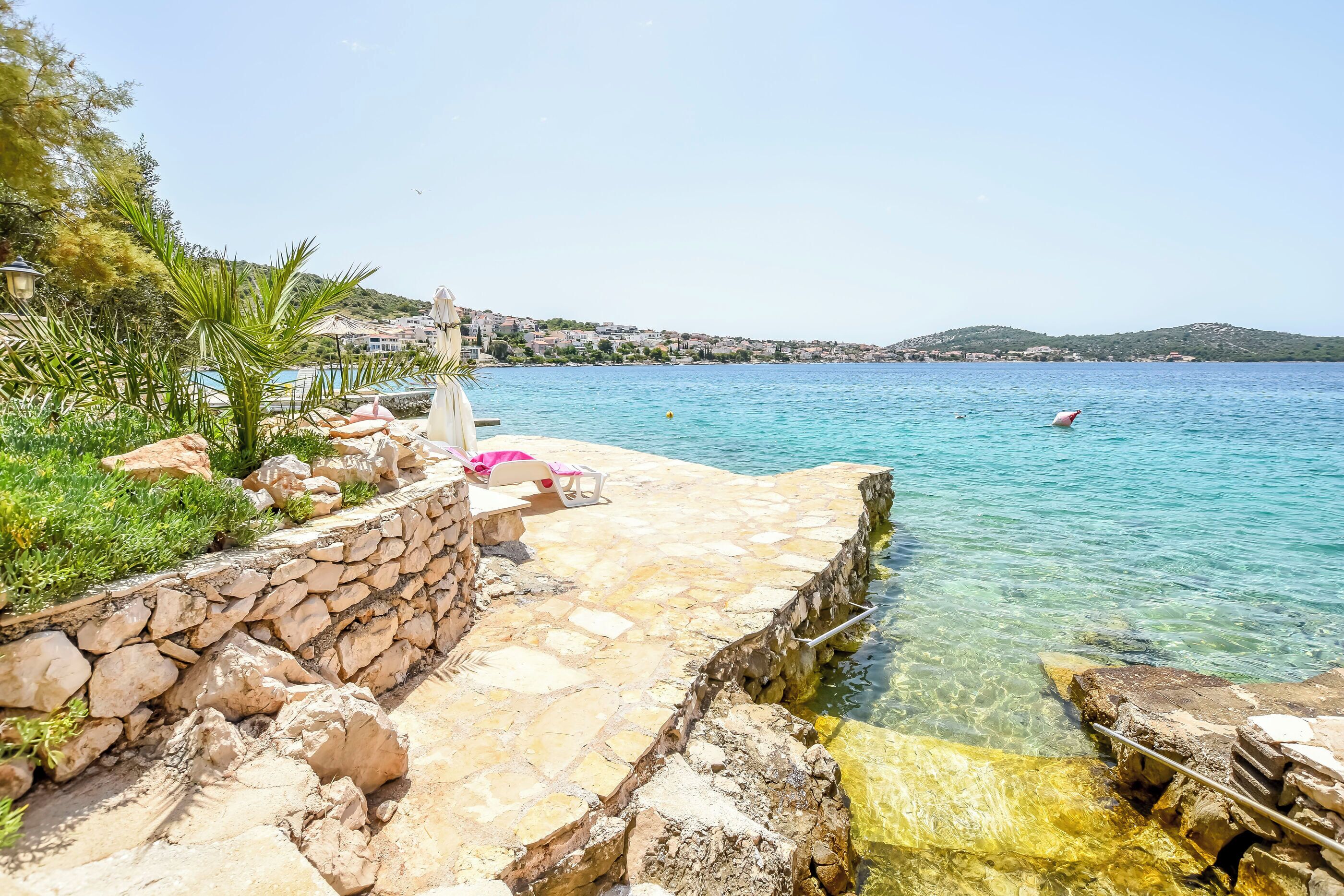A stone-paved waterfront with clear water, a pool, and a view of a hillside town.