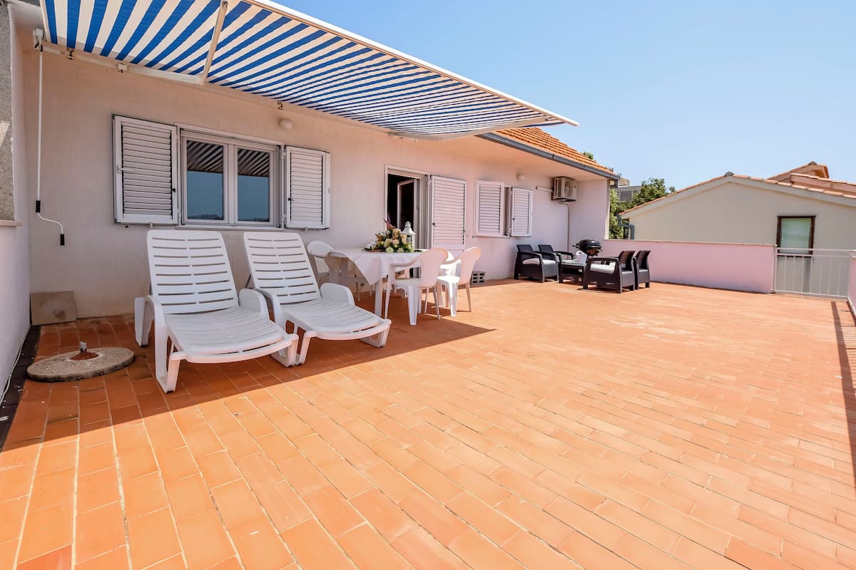 Un patio techado con dos sillones blancos, una mesita con un florero y un juego de parrilla.