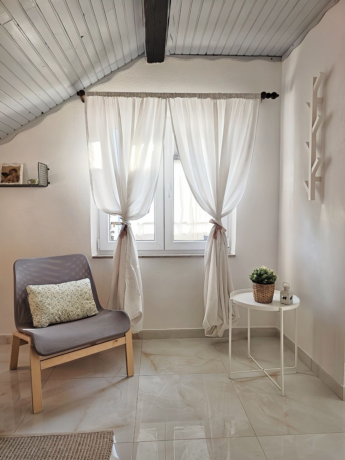 A room with a wooden ceiling, a window with white curtains, a grey chair with a cushion, a small white table with a plant and a lantern, and a wall-mounted shelf with a picture frame.