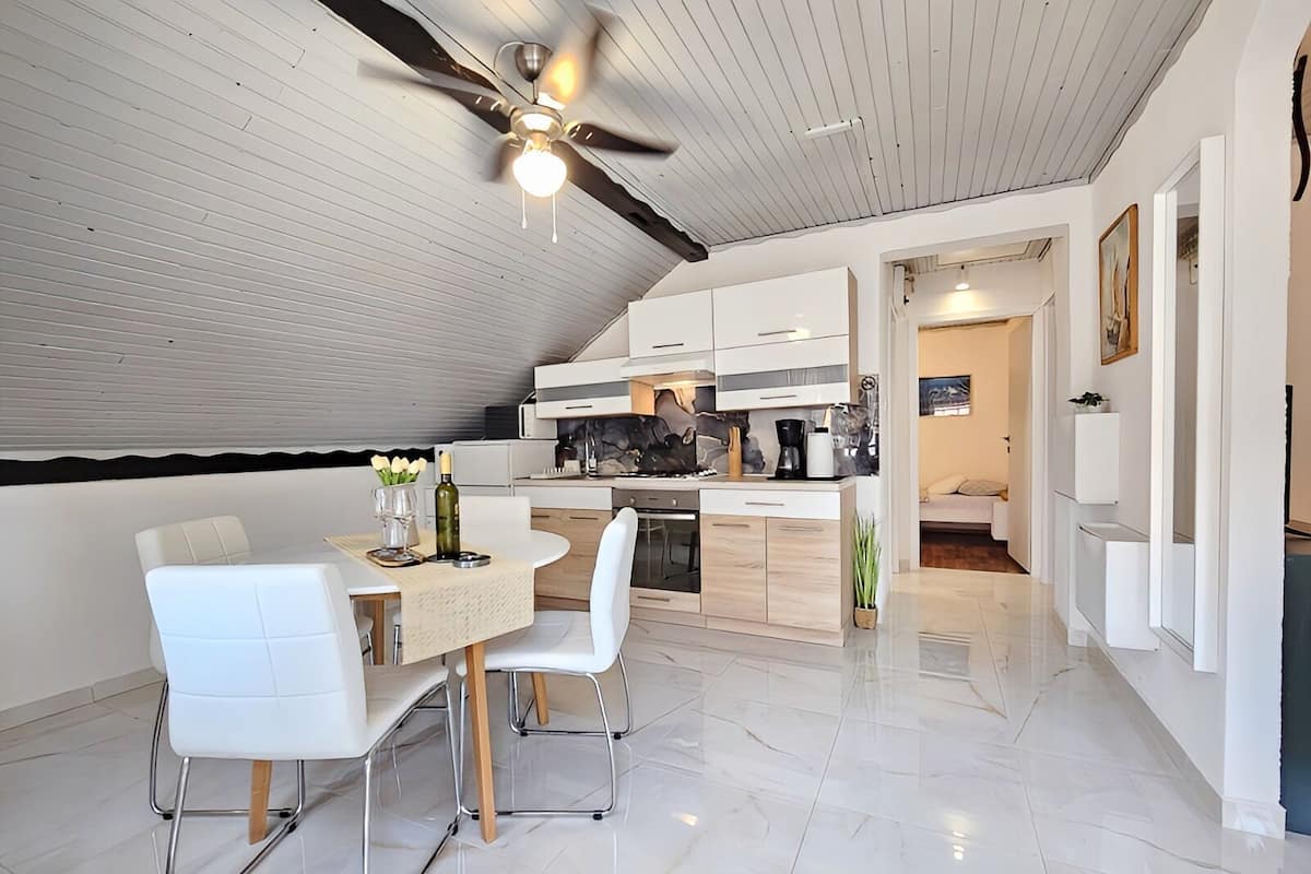 Une cuisine moderne avec des meubles blancs, un coin repas doté d’une table ronde et de chaises blanches, et un ventilateur de plafond.