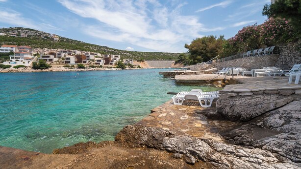 A coastal area with clear turquoise water, a rocky shoreline, and a row of white lounge chairs along the edge. - Apartments Ivica (Bilo)