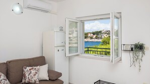 A room with a white air conditioner, a brown sofa with floral cushions, a white cabinet, and a window with a view of a coastal town. - Apartments Ivica (Bilo)
