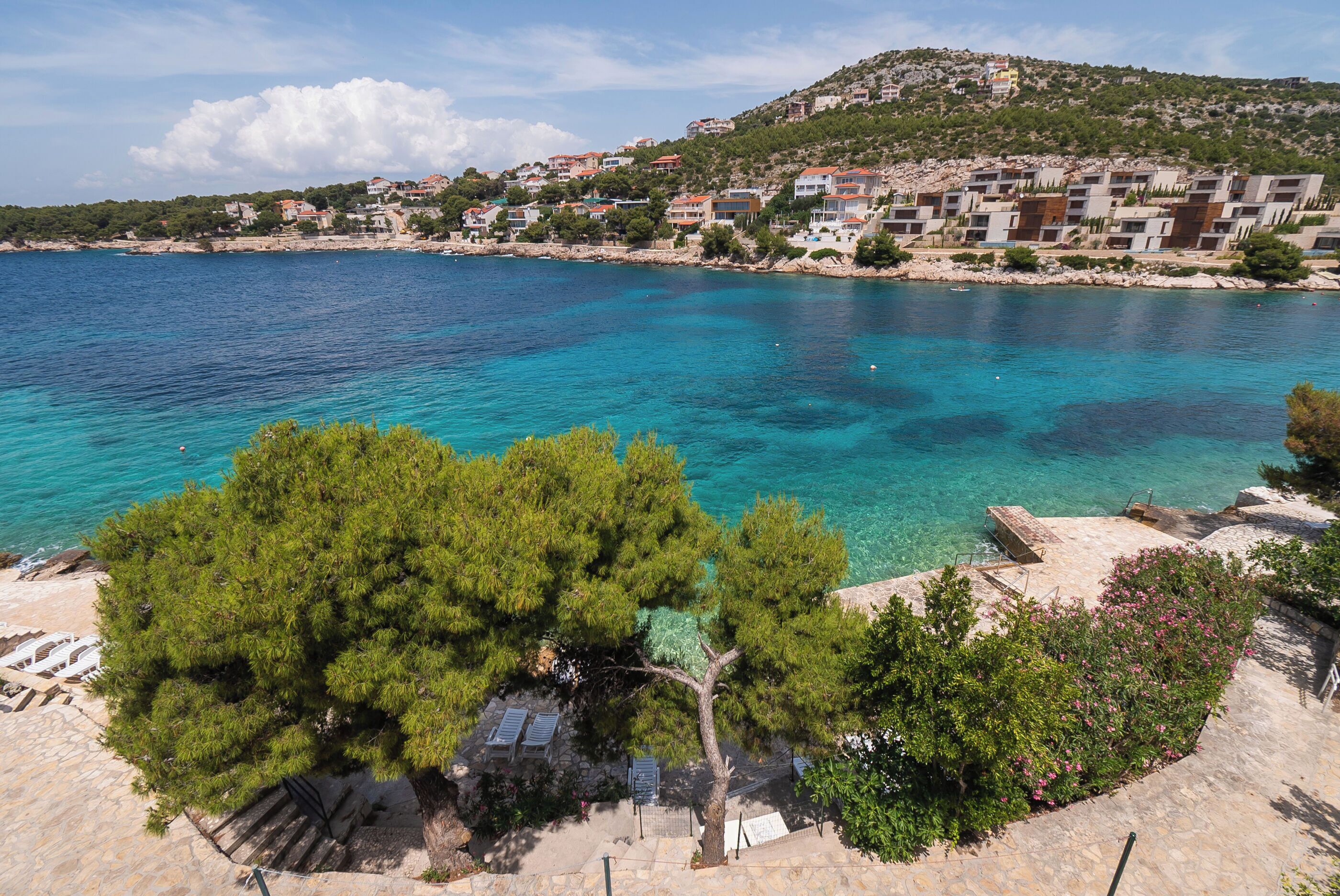 Una zona costera con aguas azules cristalinas, una escalera de piedra que desciende a la playa y una colina con edificios.