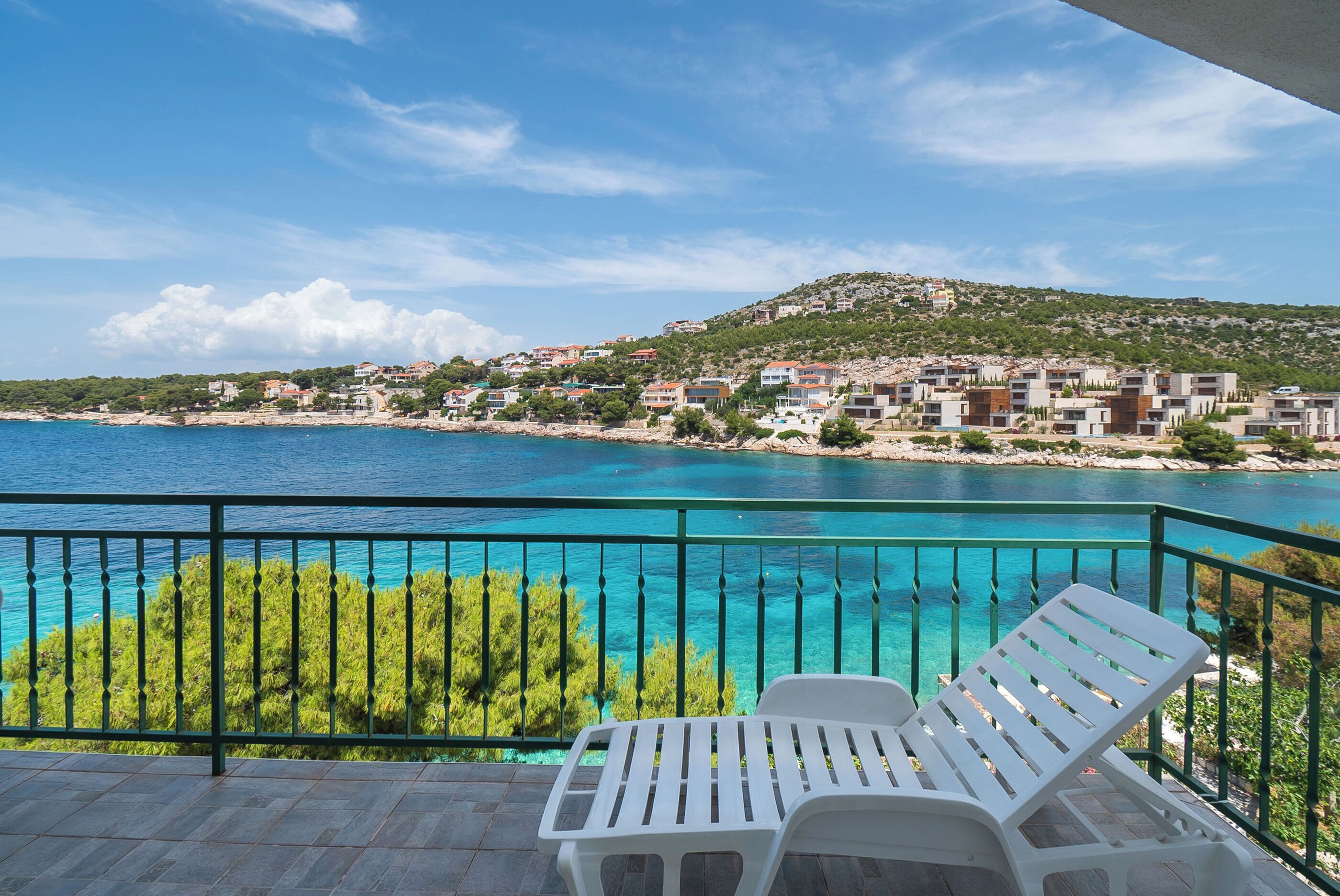 A balcony with a view of a coastal town, clear blue sea, and green vegetation.