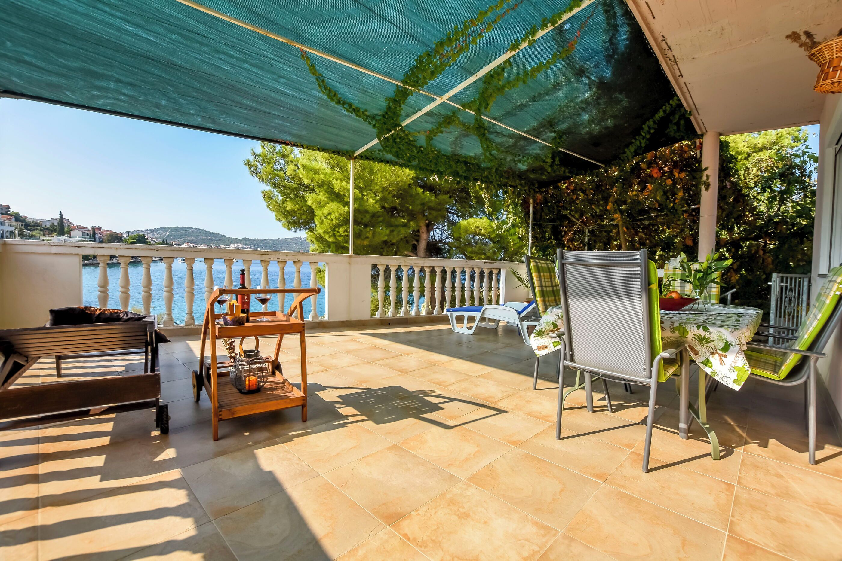A covered outdoor seating area with a view of the sea, a wooden table, a bench, and a chair.