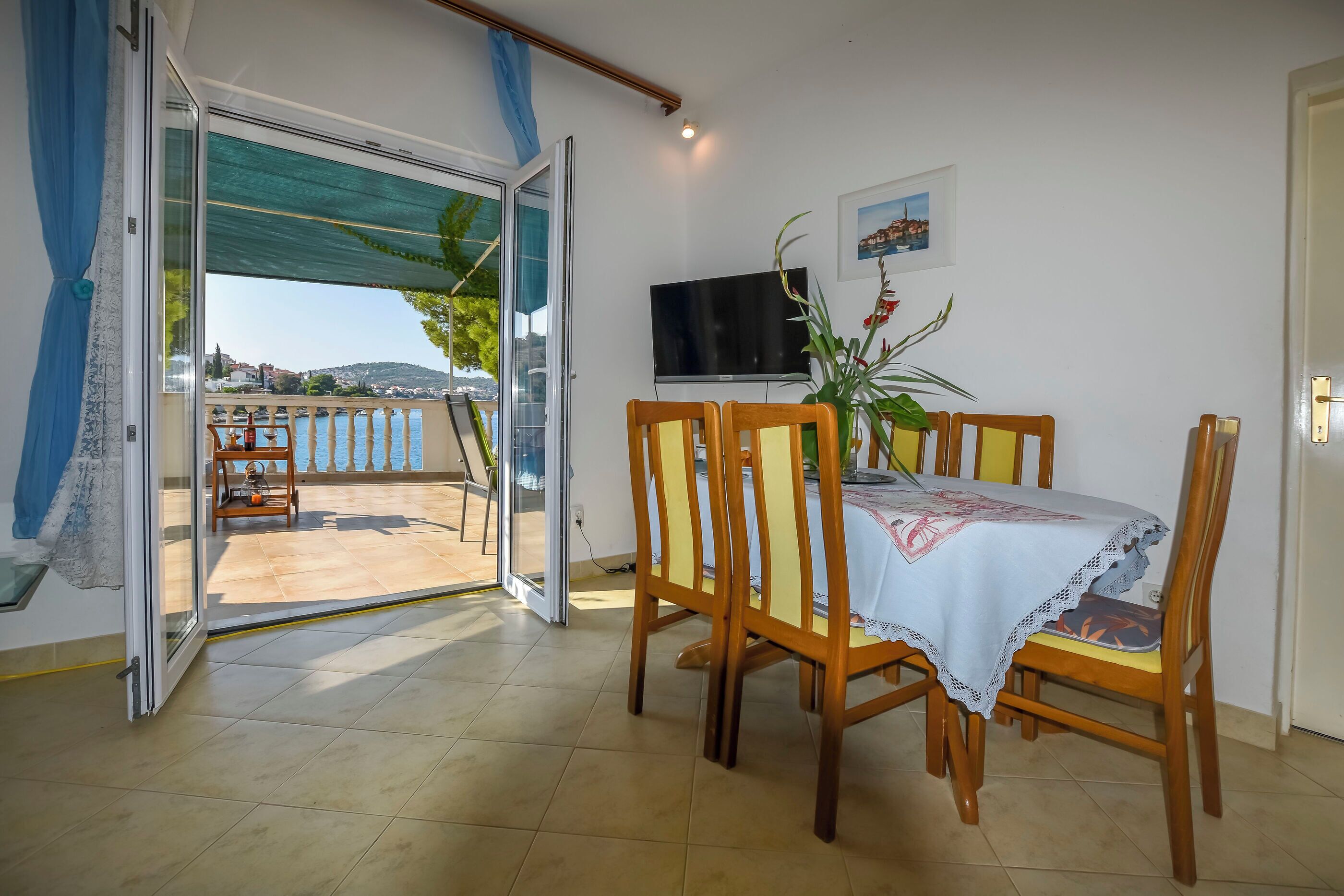 A dining area with a table and chairs, a TV on the wall, and a view of a body of water through an open door.