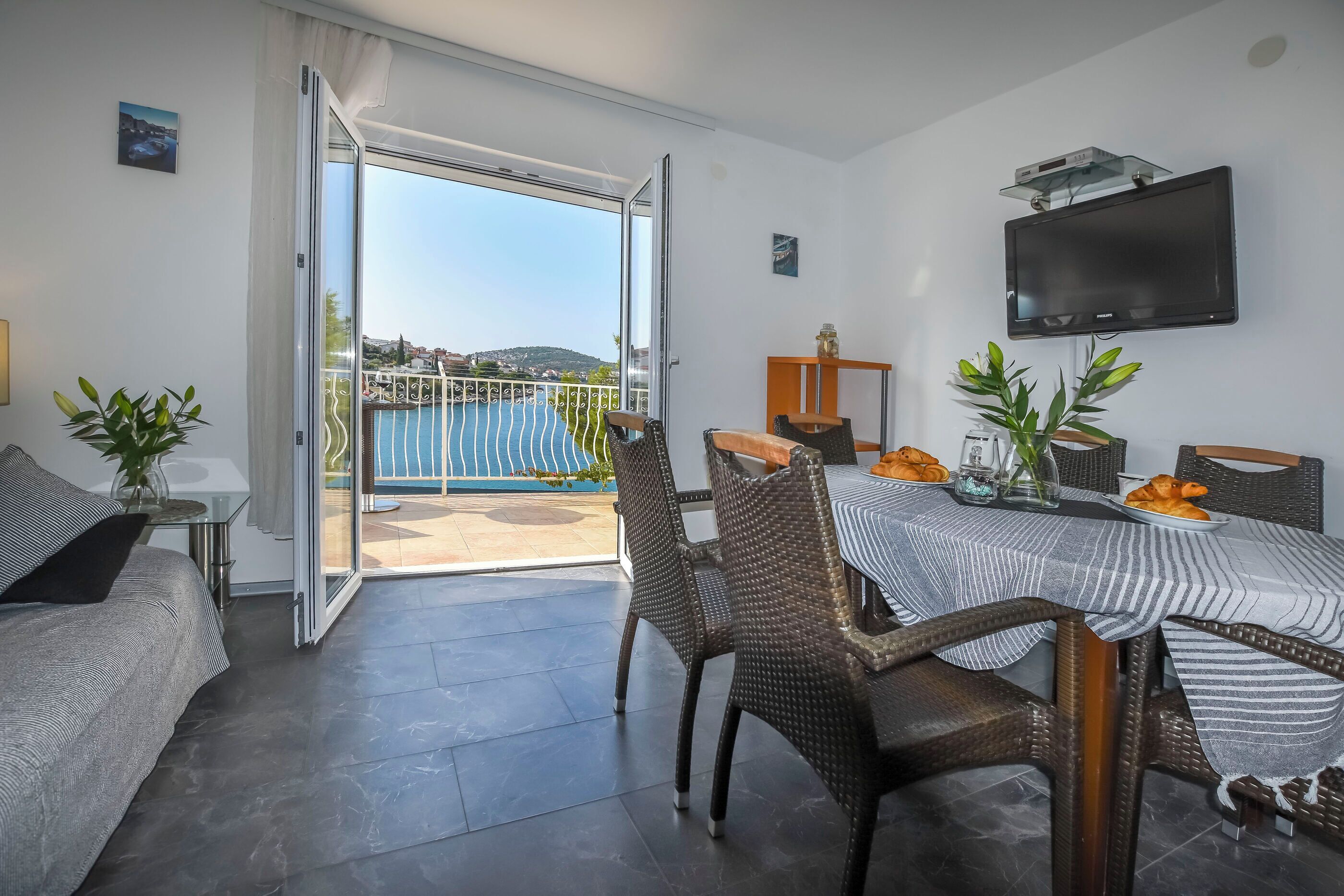 A modern living room with a dining area, a TV mounted on the wall, and a balcony with a view of the sea.