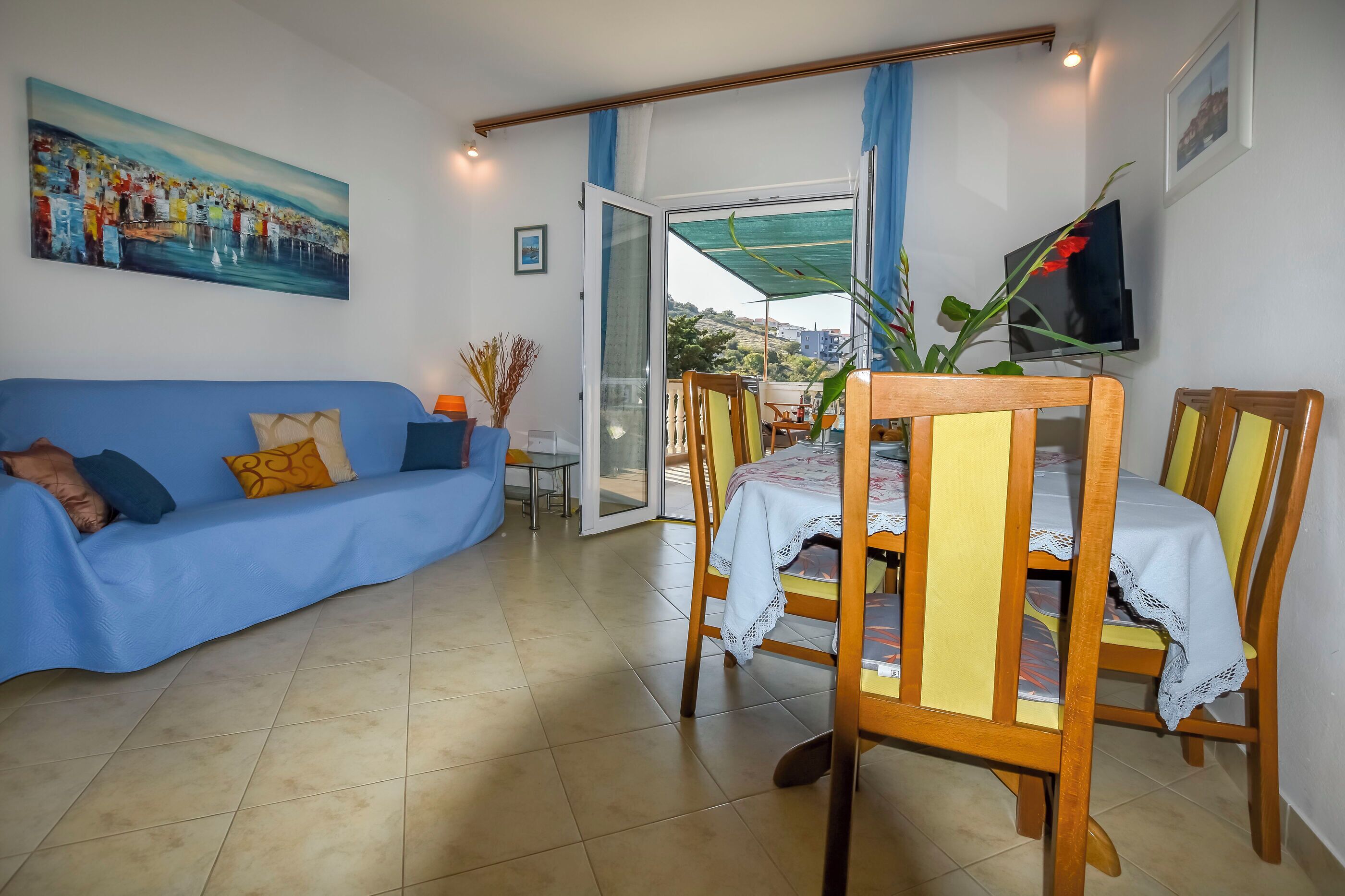 Un salón con un sofá azul, sillas de comedor de madera, una mesa con mantel de encaje, un cuadro en la pared y un televisor sobre un mueble.