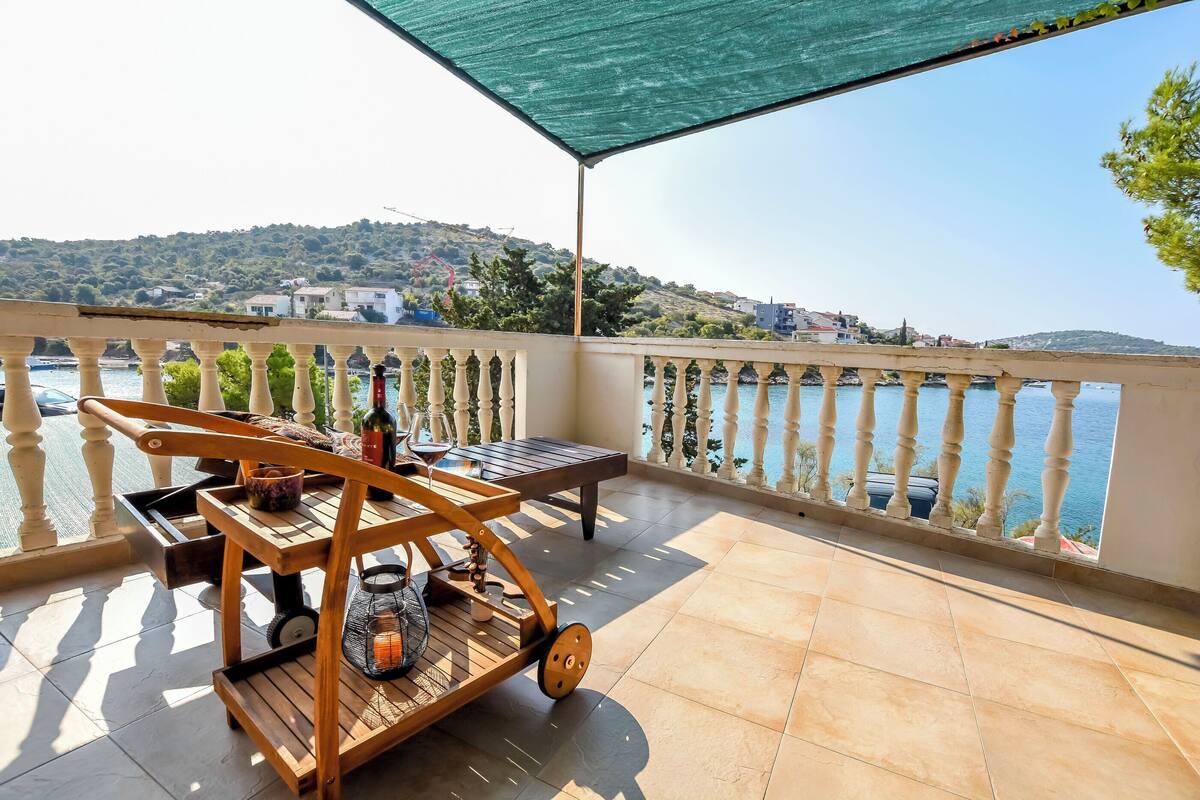 Ein Balkon mit Meerblick, ein Holzwagen mit einer Flasche und Kerze sowie ein Tisch mit einem Stuhl.