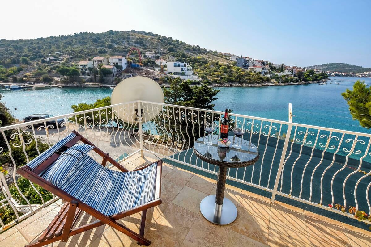 Ein Balkon mit Stuhl, Tisch und Weingläsern, sowie Blick auf ein Gewässer und Gebäude in der Ferne.