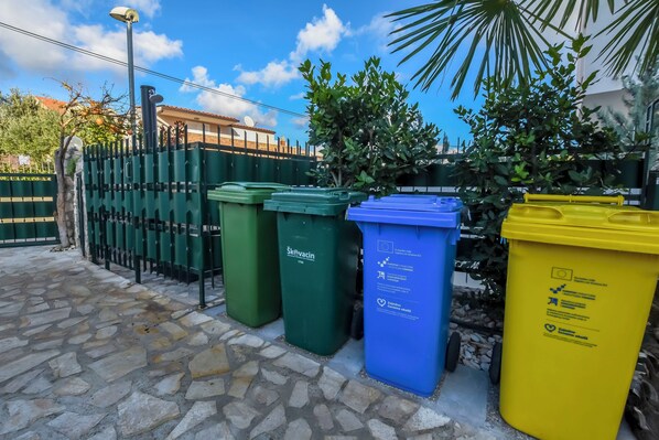A row of green, blue, and yellow recycling bins lined up against a fence with a stone-paved ground.