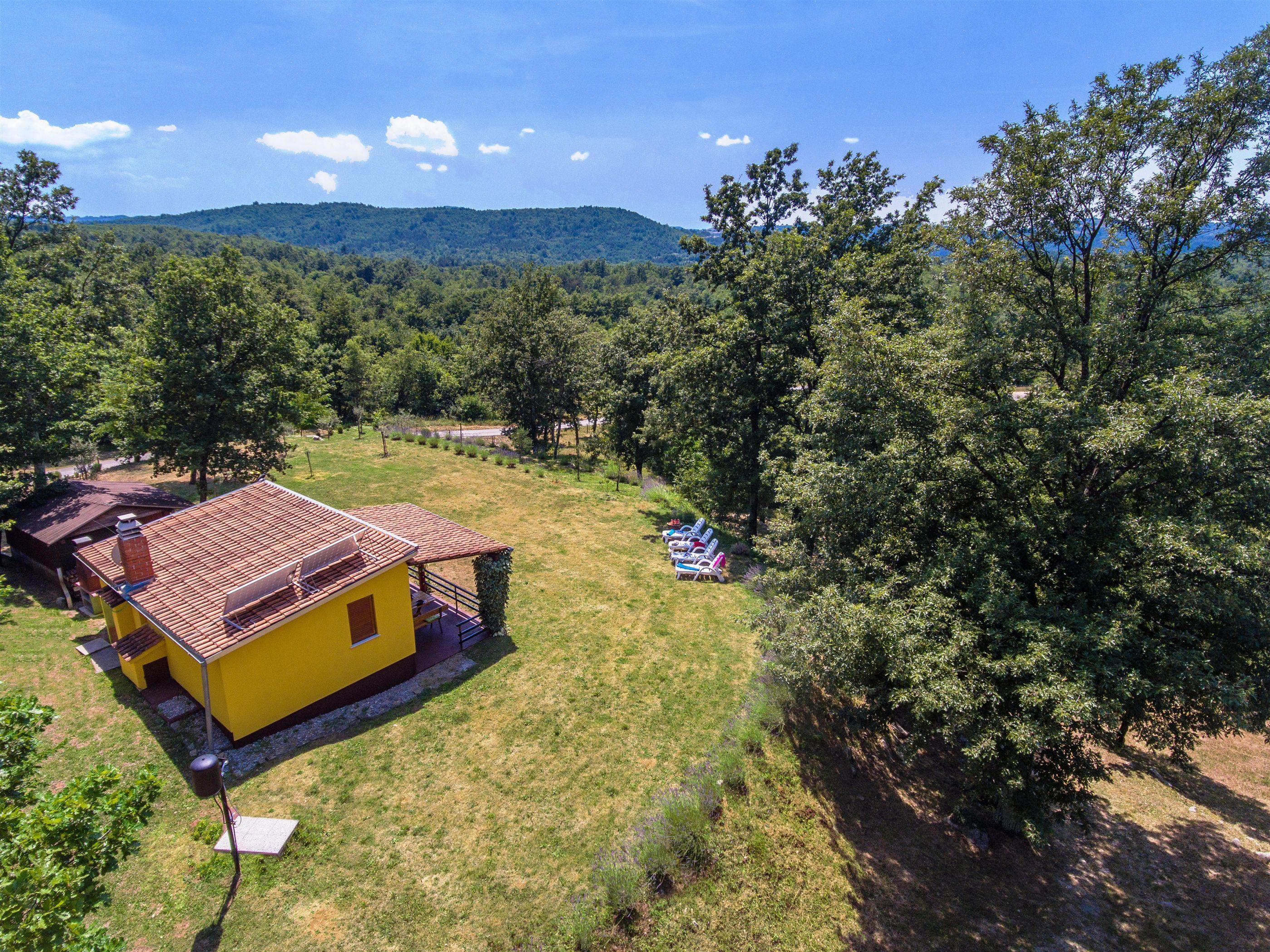 A yellow building with a red roof, surrounded by a grassy area and trees, with mountains in the background.