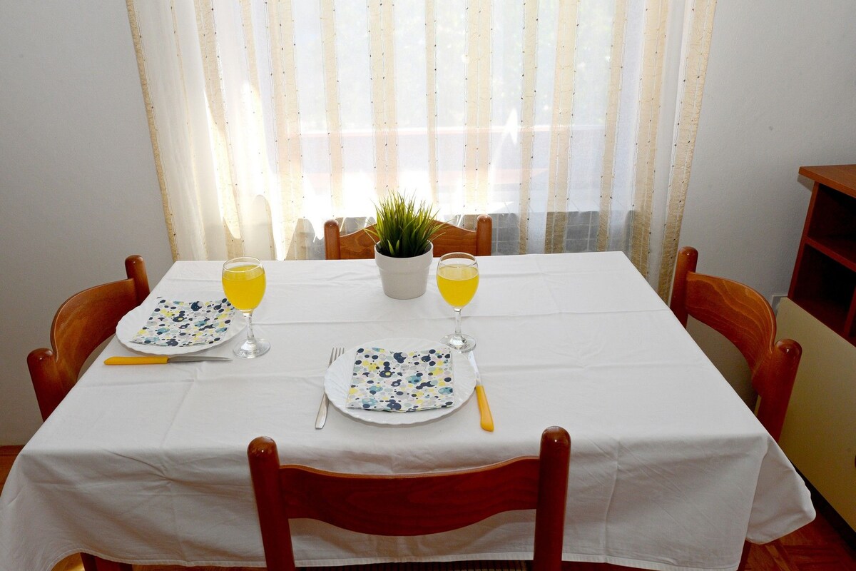 A dining table with a white tablecloth, two plates with cutlery, and two glasses of orange juice. There is a potted plant on the table, and the room has wooden chairs.