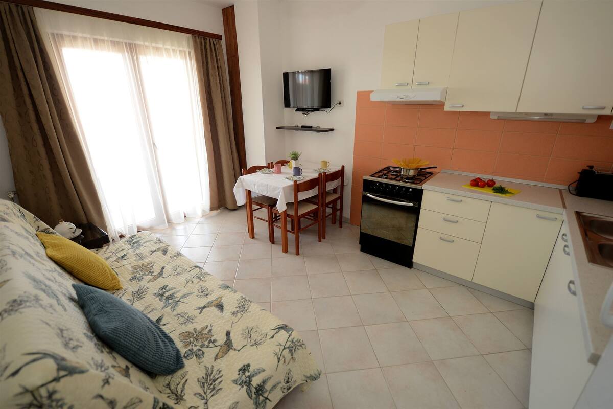 A compact living space with a sofa, a dining table, a TV mounted on the wall, a kitchen area with a stove and cabinets, and a window with curtains.
