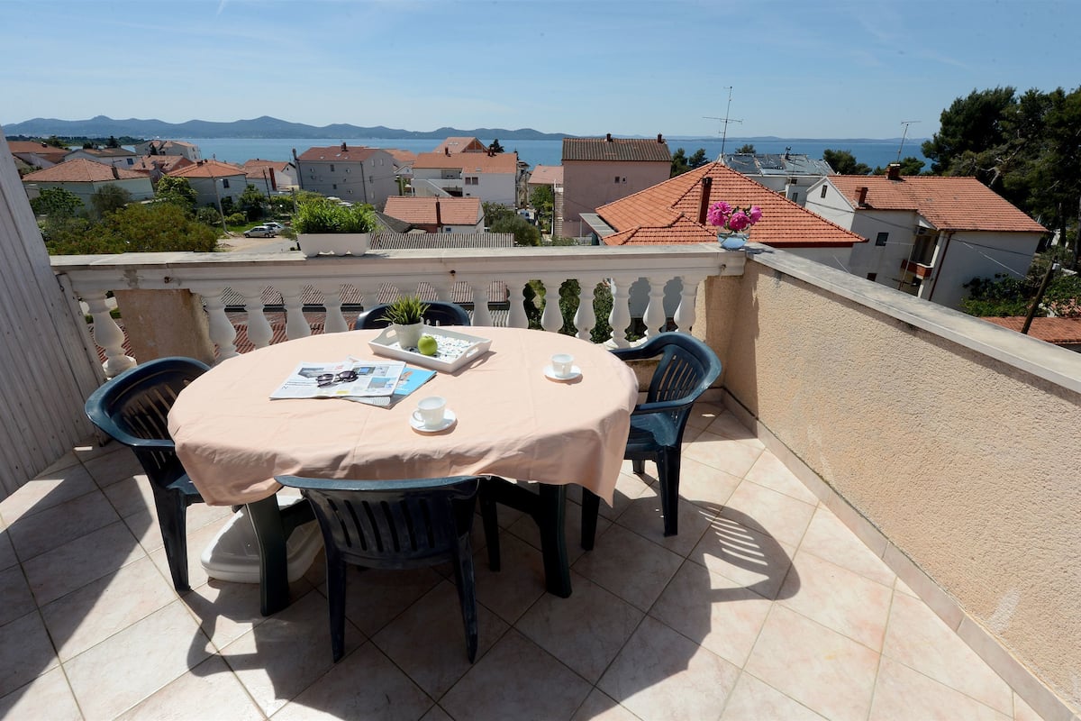 A balcony with a table set for two, overlooking a coastal town with houses and a view of the sea.