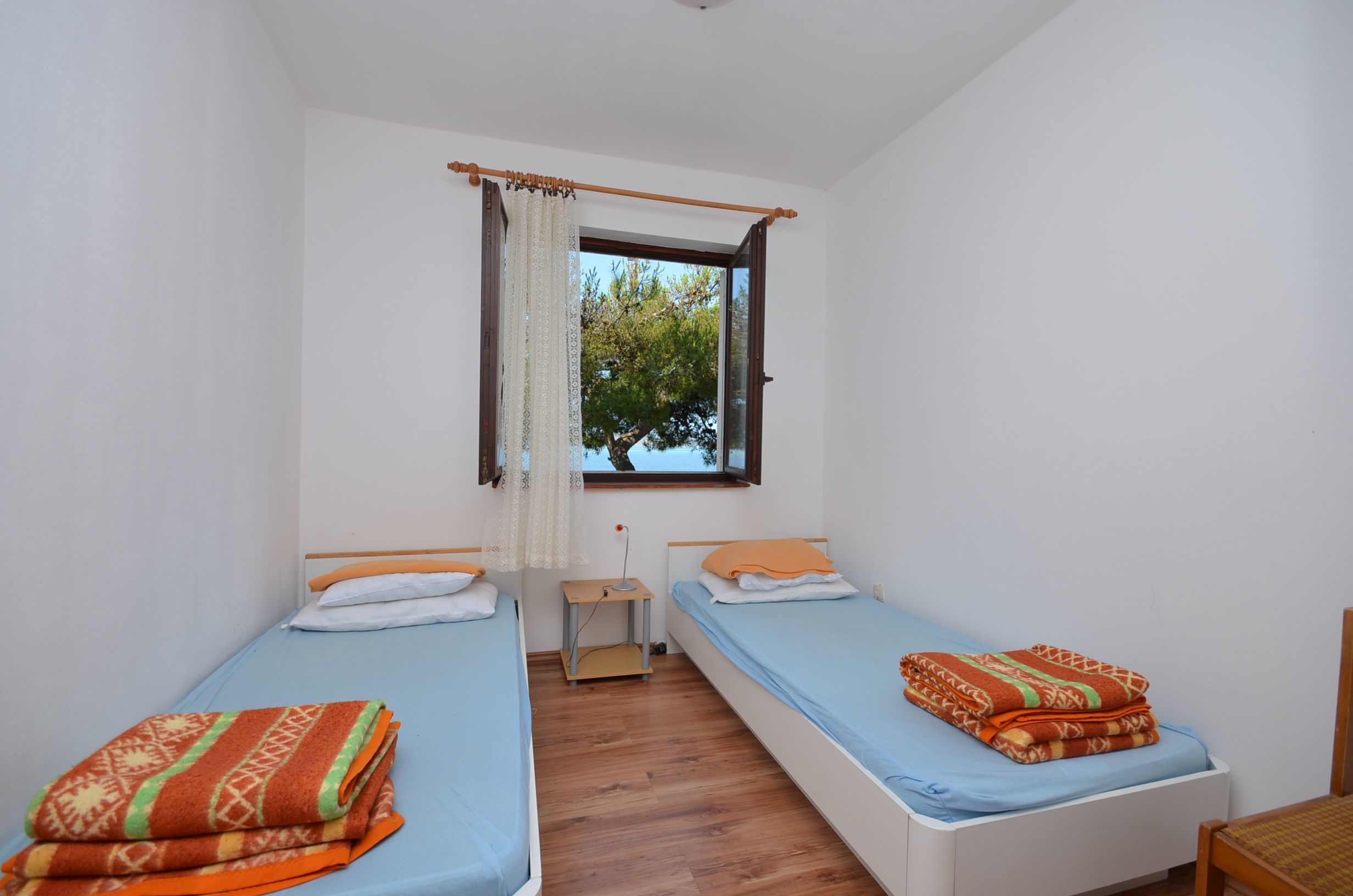 A small, clean room with two single beds, a wooden nightstand, a window with curtains, and a view of trees outside.