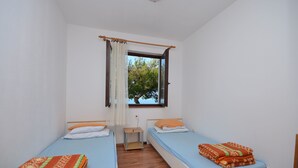 Une petite chambre propre, avec deux lits simples, une table de chevet en bois, une fenêtre avec des rideaux et une vue sur les arbres à l’extérieur.