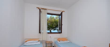 A small, clean room with two single beds, a wooden nightstand, a window with curtains, and a view of trees outside.