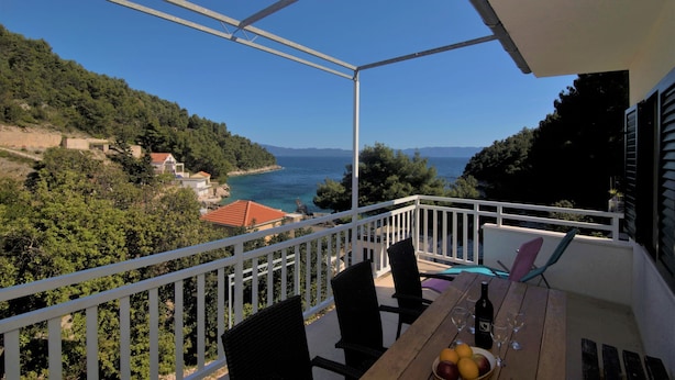 A balcony with a view of the sea and hills, wooden furniture, and a table with a bottle and glasses. - Apartment Antonio (Bogomolje)