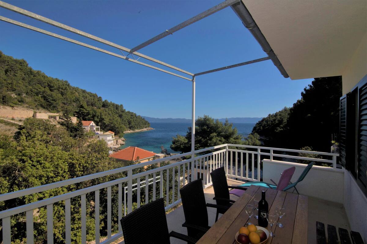 A balcony with a view of the sea and hills, wooden furniture, and a table with a bottle and glasses.