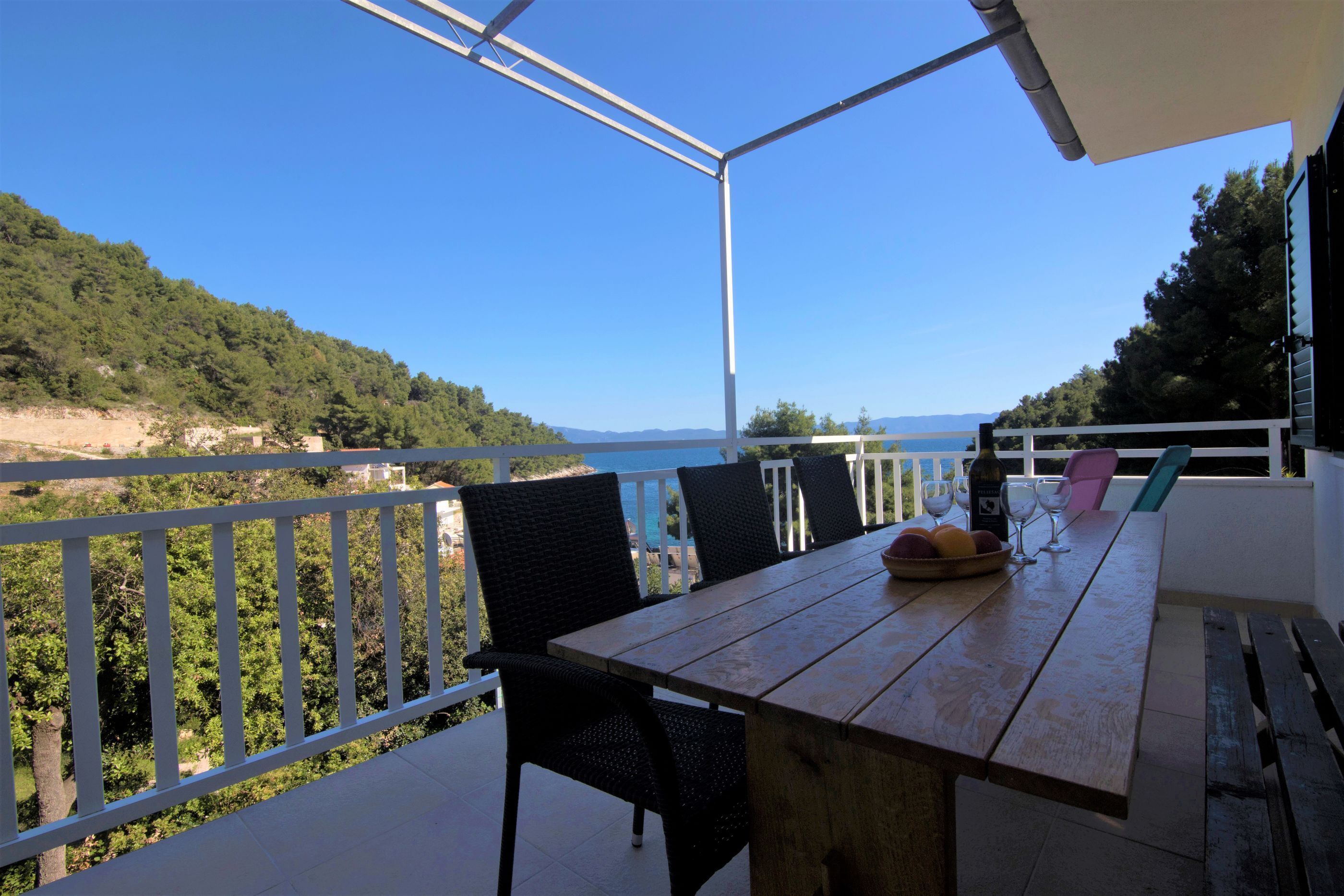 Mesa de madeira com cadeiras numa varanda com vista panorâmica para uma praia e montanhas.