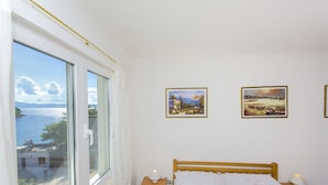 A bedroom with a bed, two bedside tables, a window with a view of the sea, and two framed pictures on the wall. - Apartment Antonio (Bogomolje)