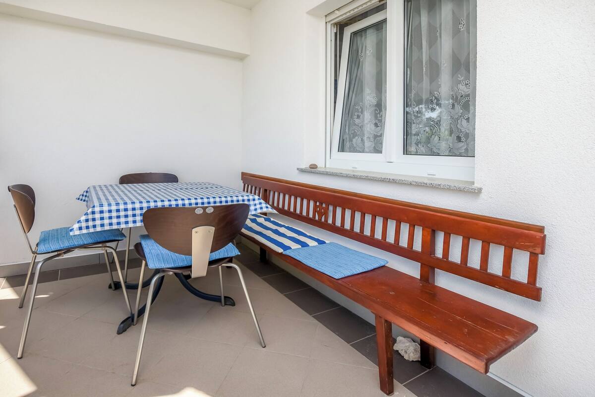 A small outdoor dining area with a table and chairs, a wooden bench, and a window with curtains.