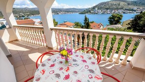 A balcony with a table set for two, a vase with flowers, and a view of a lake and town. - Apartments Sopic (Rogoznica)