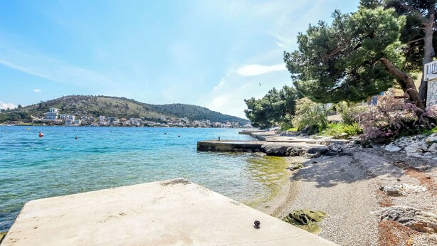 A coastal scene with a clear blue sea, a concrete pier, rocky shore, and a hill with buildings in the background. - Apartments Sopic (Rogoznica)