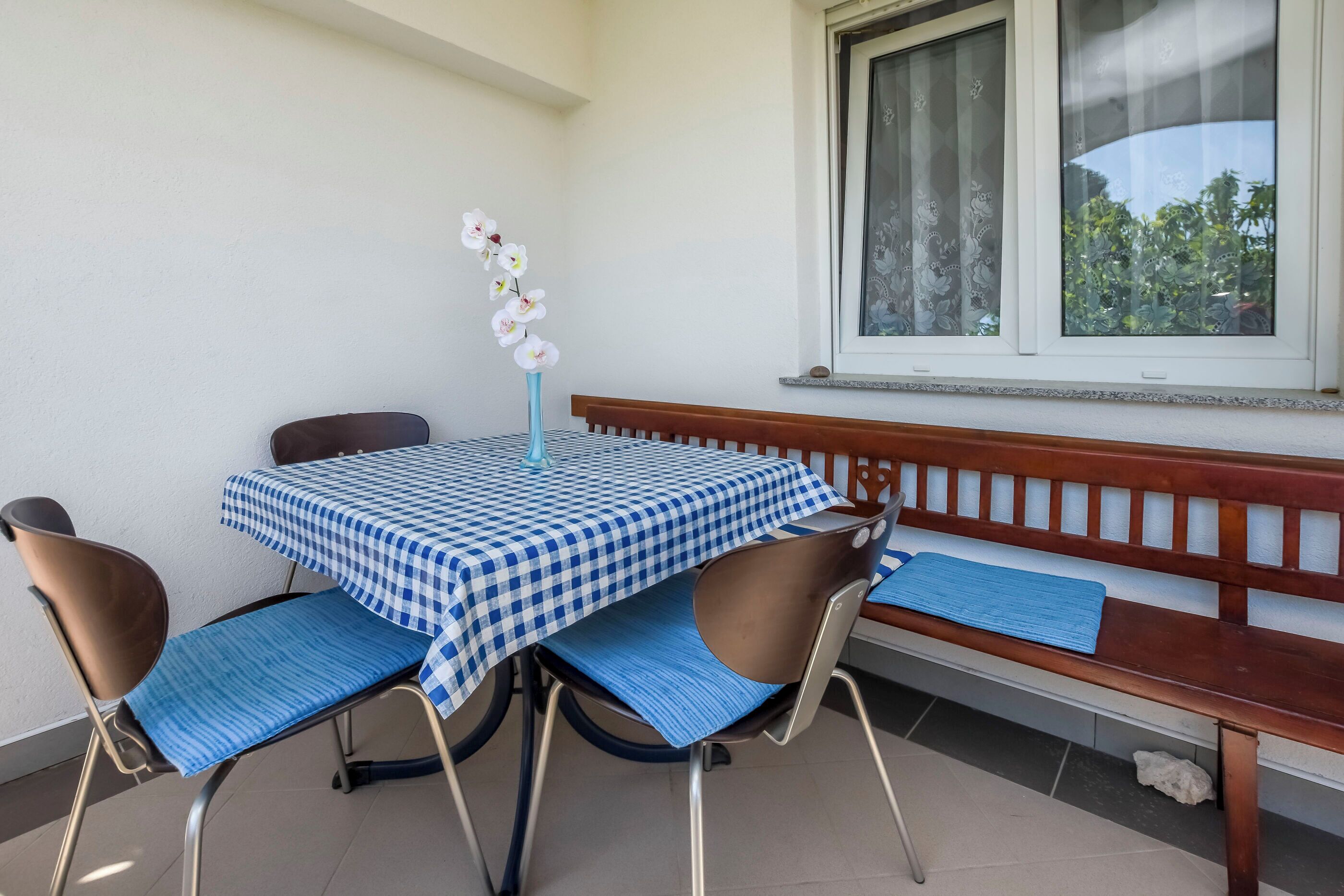 A balcony with a table and chairs, a bench, and a window with a view of trees.